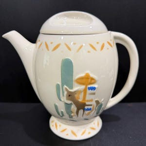 May include: Cream-colored ceramic teapot with a lid, spout, and handle. The teapot features a Southwestern design with a cactus, donkey, and decorative accents in orange and teal. The teapot sits on a small pedestal base.
