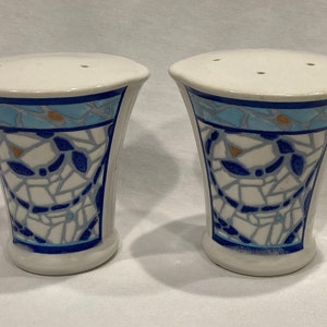 May include: A pair of white ceramic salt and pepper shakers. Each shaker has a flared, vase-like shape with a mosaic-style design in blue, white, and gray. The top rim features a blue band, and the top has three holes.
