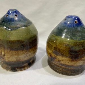 Puede incluir: Dos saleros y pimenteros de cerámica con un esmalte azul, verde y marrón. Los saleros tienen una forma redondeada y un pequeño agujero en la parte superior para dispensar sal y pimienta.