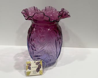 May include: A purple glass vase with a ruffled rim and a molded floral pattern on the exterior. A square tag is attached to the base, featuring illustrations of mulberries and the text Mulberry, Created Especially for you by Fenton.