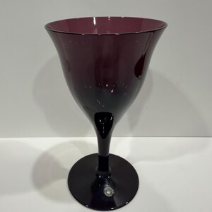 May include: A deep purple glass goblet with a wide, flared bowl and a smooth, solid stem that transitions into a circular base. The glass features a translucent, dark plum hue throughout, with a small circular sticker visible on the base.
