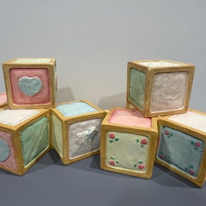 May include: A set of eight ceramic blocks with different pastel colored designs. Each block has a different design, including hearts, flowers, and a lamb. The blocks are stacked in a pyramid shape.