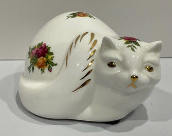 May include: A white ceramic cat figurine with gold accents and floral decals. The cat is curled up, with gold details on its side and floral designs on its head and body. The eyes and nose are also gold.