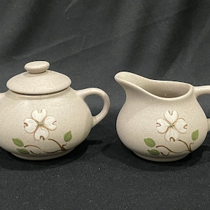 May include: A set of two ceramic sugar and creamer containers with a speckled beige glaze. Each piece features a white flower with green leaves painted on the front.
