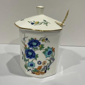 May include: White ceramic honey pot with a gold trim and floral design featuring blue, yellow, and green flowers. The pot has a lid with a small spoon attached.