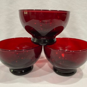 May include: Three ruby red glass bowls with a speckled texture. Each bowl has a rounded shape and sits on a black pedestal base. The bowls are arranged in a stacked formation, showcasing their design.