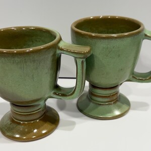 May include: Two green ceramic mugs with a pedestal base and a handle. The mugs have a rounded bowl shape and a brown rim. The handles are rectangular. The mugs are a muted green color with brown accents.