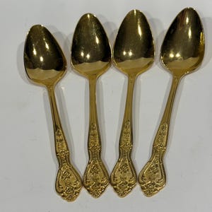 May include: A set of four gold-toned metal spoons arranged side by side. Each spoon features a polished oval bowl and a handle decorated with an intricate embossed floral pattern that extends from the neck down to the rounded base of the handle.