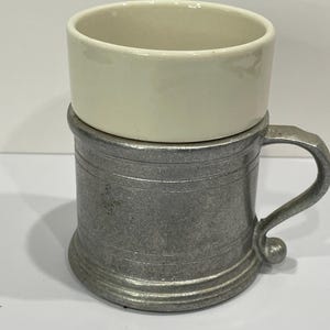 May include: A vintage shaving mug with a cream-colored ceramic bowl resting on a pewter-toned metal mug. The mug has a handle and a decorative, ridged design around the base and body. The items are likely from the early to mid-20th century.