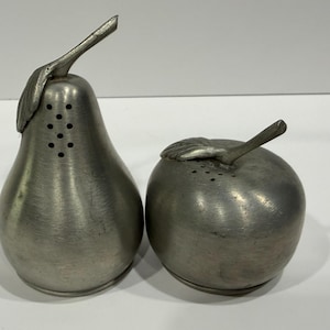 May include: A pair of pewter salt and pepper shakers. One is pear-shaped with a leaf and stem, the other is apple-shaped with a hand and stem. Both have small holes for dispensing.