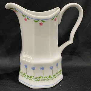 May include: White ceramic pitcher with a curved handle and a spout. The pitcher is decorated with blue and pink floral patterns and green vines. A blue line borders the top and bottom of the pitcher.