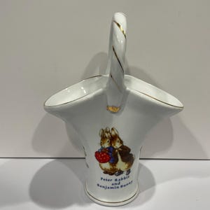 May include: White porcelain basket with a gold handle and a picture of Peter Rabbit and Benjamin Bunny. The basket is decorated with blue and red accents.