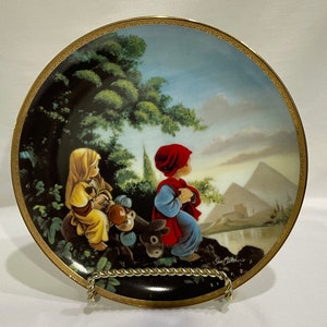 May include: A decorative porcelain plate with a gold rim features a painting of two children in a landscape setting. The children are wearing red and yellow clothing and are sitting on a rock with a small animal. The background includes a green tree, a blue sky, and a distant mountain range. The plate is signed "Sue Walters" in the lower right corner.