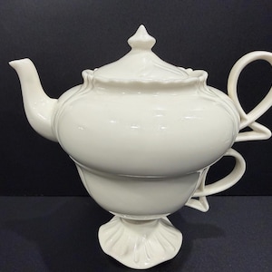 May include: Cream-colored ceramic teapot and teacup set. The teapot has a rounded body, a curved spout, and a lid with a finial. The teacup is integrated into the base of the teapot, with a handle. The set is on a dark surface.