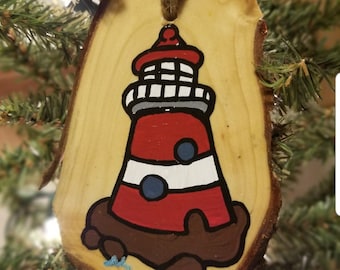 May include: A hand-painted wooden ornament shaped like a lighthouse. The lighthouse is red and white with a grey roof and blue details. The ornament is hanging on a Christmas tree.