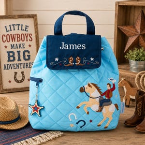 May include: A light blue quilted backpack featuring a dark blue flap with the name James embroidered in white, accented by cowboy boot motifs. The front displays an embroidered cowboy riding a horse, a horseshoe, and a star-shaped zipper pull.