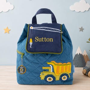 May include: A quilted blue backpack featuring a dark blue flap with an embroidered crane and the name Sutton in yellow. The front displays a yellow and green dump truck applique and a circular tag with the text Let's Roll on a zipper pull.