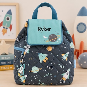 May include: A quilted dark navy blue backpack featuring a space-themed pattern of rockets, planets, and stars. The top flap is light blue with the name Ryker embroidered in black. It includes a top handle, a front zipper pocket, and a rocket charm.