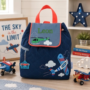 May include: A navy blue quilted backpack featuring a top flap with the embroidered name Leon, a green helicopter, and white clouds. The bag includes a red handle, a front zipper pocket with a car-shaped charm, and a blue and white airplane graphic.