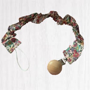 May include: A floral print pacifier clip with a wooden bead and metal clip. The fabric is a light pink with a pattern of small flowers in shades of pink, blue, and yellow. The clip is designed to attach to a baby's clothing.