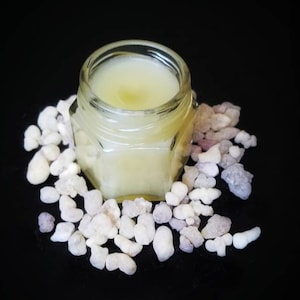May include: A small glass jar filled with a pale yellow balm, surrounded by white and grey stones on a black background.