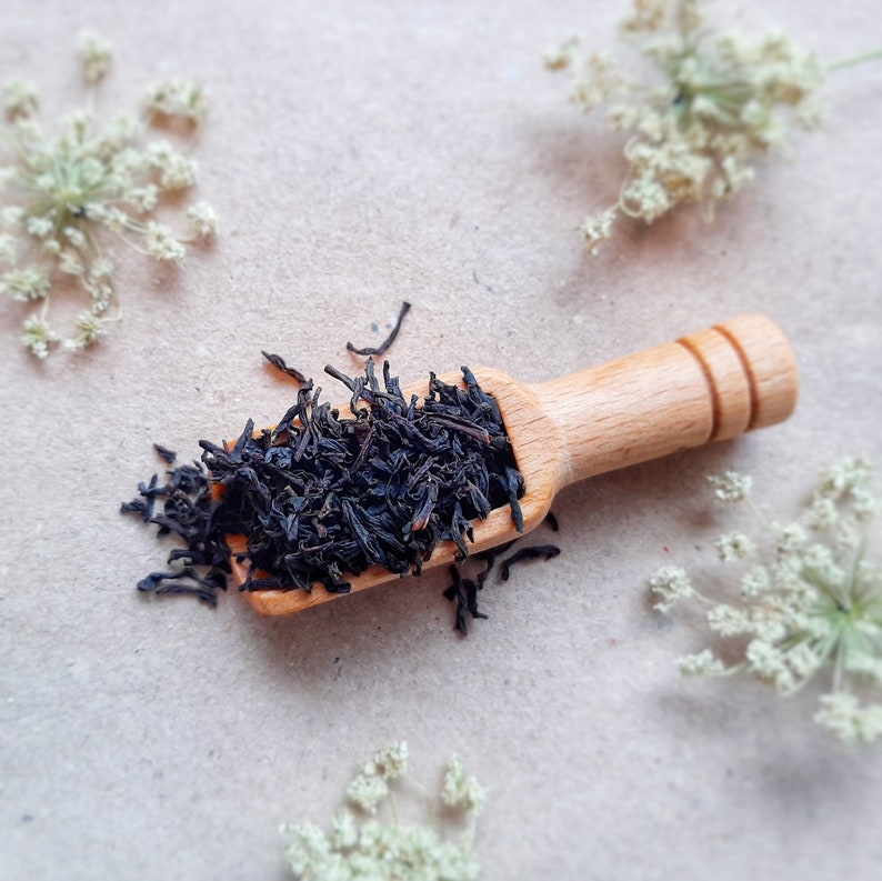 May include: A wooden scoop filled with loose leaf black tea on a light brown surface with white flowers scattered around it.