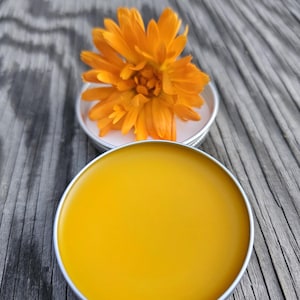 May include: A small round tin container filled with a bright yellow salve or balm. A single orange calendula flower sits on top of the tin.