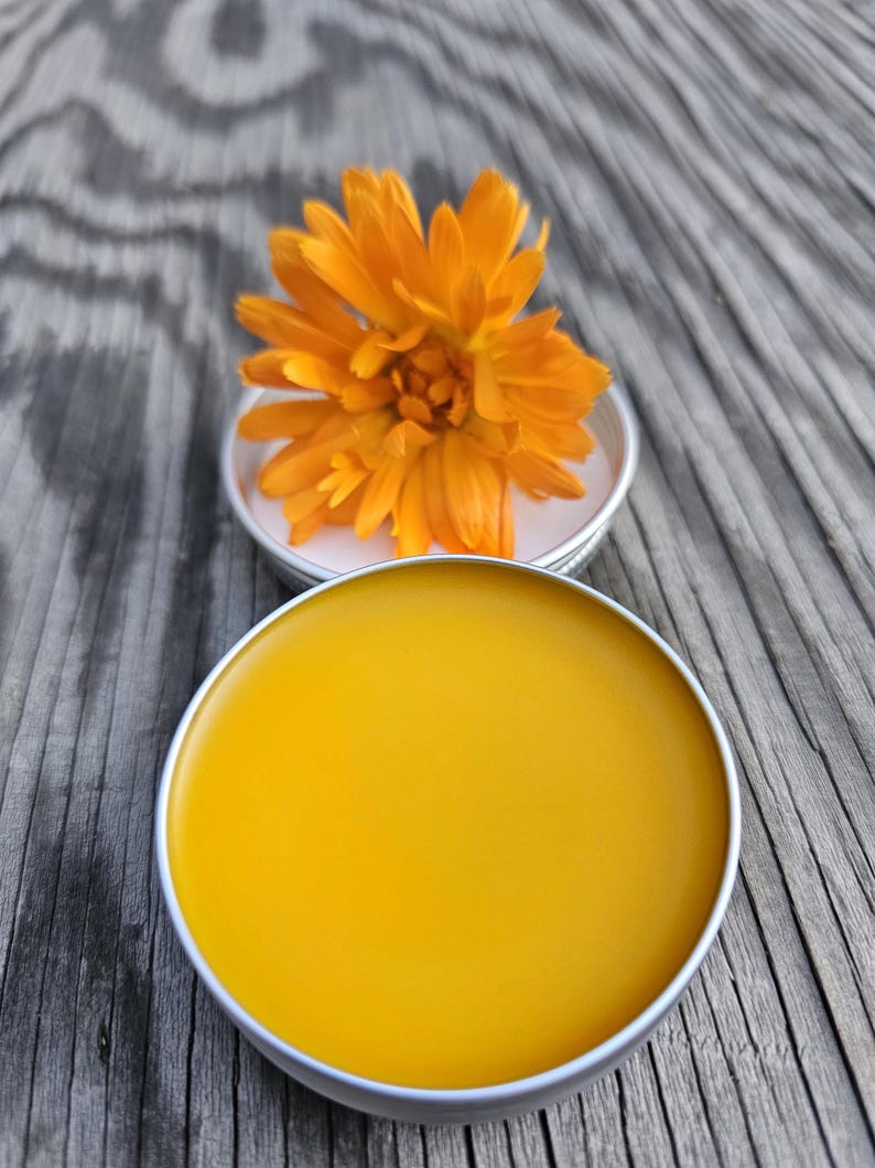 May include: A small round tin container filled with a bright yellow salve or balm. A single orange calendula flower sits on top of the tin.