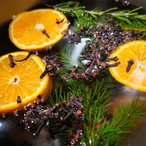 May include: A pot filled with water, orange slices, cloves, and evergreen sprigs. The ingredients are simmering to create a fragrant potpourri.