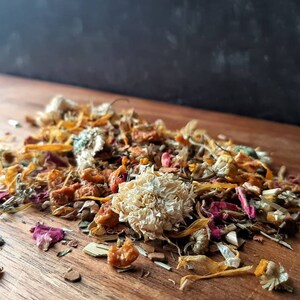 May include: A mix of dried flowers and herbs, including chamomile, rose petals, and orange peel. The mixture is a light brown color with hints of pink and yellow.