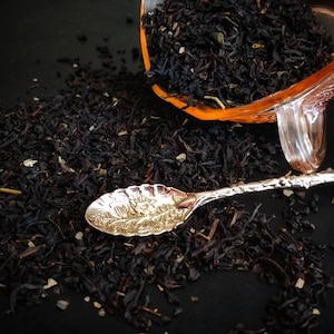 May include: A close-up of loose black tea leaves scattered on a black surface. A silver spoon with an ornate design rests on the tea leaves. A clear glass jar filled with tea leaves is overturned in the background.