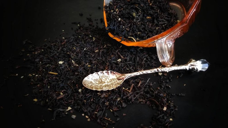 May include: A close-up of loose black tea leaves scattered on a black surface. A silver spoon with an ornate design rests on the tea leaves. A clear glass jar filled with tea leaves is overturned in the background.