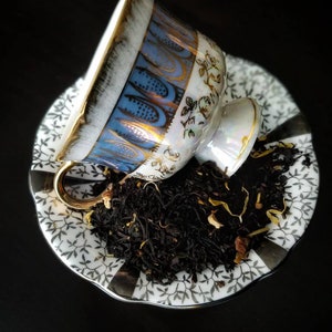 May include: A white teacup with blue and gold floral designs sits on a white saucer with a black and white floral pattern. The teacup is filled with loose leaf black tea.