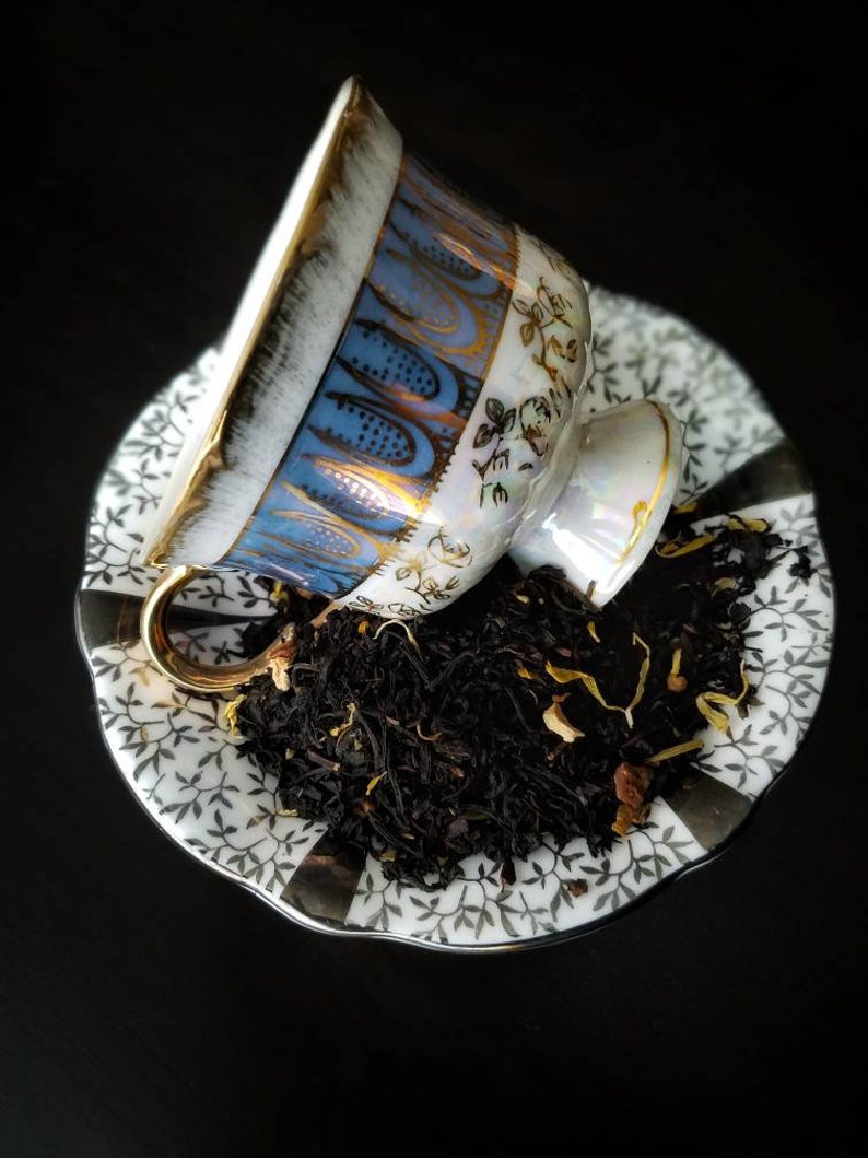May include: A white teacup with blue and gold floral designs sits on a white saucer with a black and white floral pattern. The teacup is filled with loose leaf black tea.