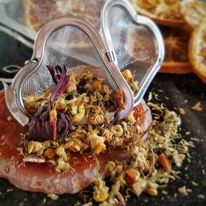 May include: A heart-shaped stainless steel tea infuser filled with dried chamomile, hibiscus, and orange peel. The infuser is sitting on a pink and white agate coaster.