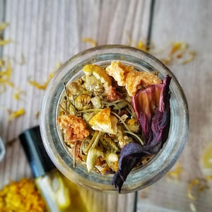 May include: A glass jar filled with a blend of dried herbs and flowers, including chamomile, hibiscus, and orange peel. The jar is sitting on a wooden surface.