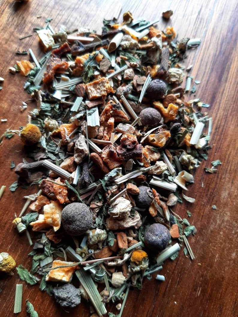 Op de afbeelding: Een close-up van een mix van gedroogde kruiden, specerijen en fruitschillen. De mix bevat groene kruiden, sinaasappelschillen, bruine kaneelstokjes en zwarte peperkorrels.