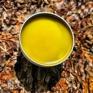 May include: A small round tin container filled with a bright yellow balm. The tin is sitting on a brown, textured surface.
