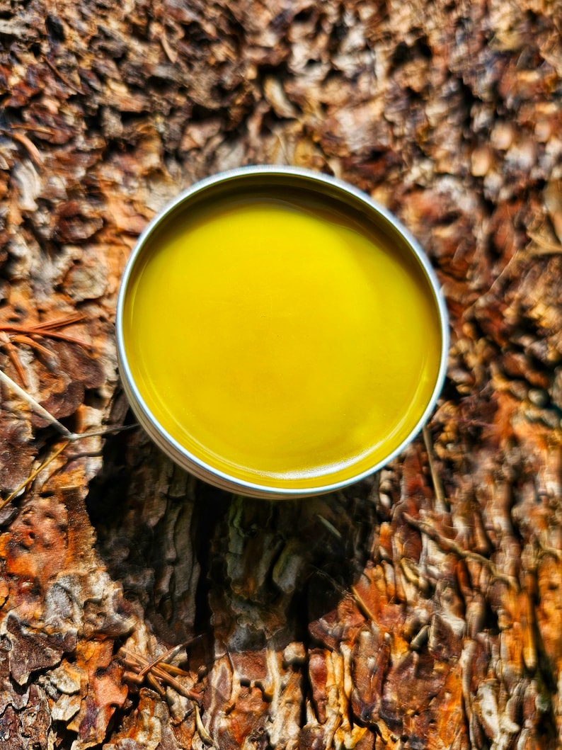 May include: A small round tin container filled with a bright yellow balm. The tin is sitting on a brown, textured surface.