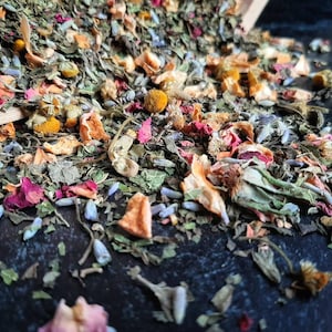 Op de afbeelding: Een mix van gedroogde kruiden, bloemen en fruitstukjes. De mix bevat groene kruiden, roze en rode bloemblaadjes, oranje fruitstukjes en gele bloemknoppen.