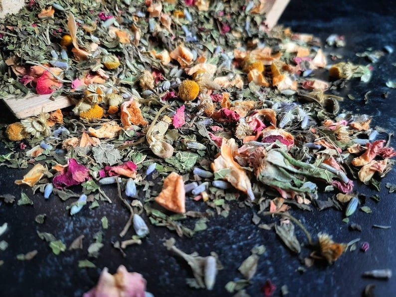 Op de afbeelding: Een mix van gedroogde kruiden, bloemen en fruitstukjes. De mix bevat groene kruiden, roze en rode bloemblaadjes, oranje fruitstukjes en gele bloemknoppen.