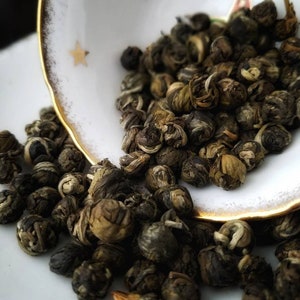May include: Close-up of green tea pearls, also known as jasmine pearls, in a white teacup with gold trim. The pearls are small and round, and they are arranged in a pile.