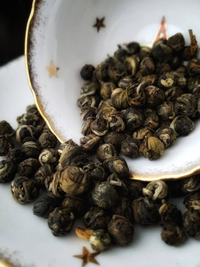 May include: Close-up of green tea pearls, also known as jasmine pearls, in a white teacup with gold trim. The pearls are small and round, and they are arranged in a pile.