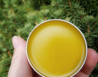 Puede incluir: Un pequeño recipiente redondo de hojalata lleno de un bálsamo amarillo brillante. El recipiente es de metal y tiene una tapa plateada.