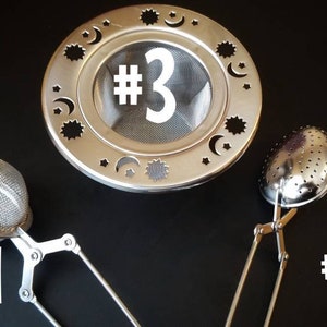 Op de afbeelding: Drie verschillende thee-infusers. De eerste is een ronde bal-infuser met een handvat. De tweede is een ronde infuser met een handvat en een veerbelaste opening. De derde is een ronde, platte infuser met een handvat en een decoratief ontwerp van sterren en manen.
