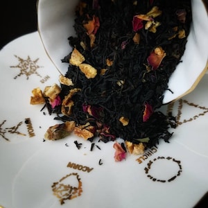 May include: A white porcelain teacup with a gold rim is tipped over, spilling black tea leaves, orange peel, and pink rose petals onto a white saucer with gold designs. The saucer has the words "Circle", "Woman", "Acorn", and "Sun" printed on it.