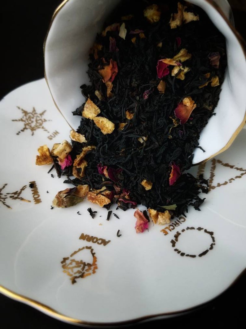 May include: A white porcelain teacup with a gold rim is tipped over, spilling black tea leaves, orange peel, and pink rose petals onto a white saucer with gold designs. The saucer has the words "Circle", "Woman", "Acorn", and "Sun" printed on it.