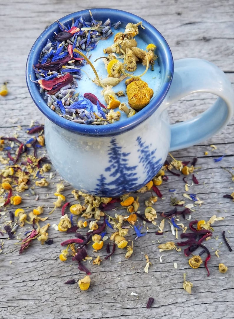Pode incluir: Uma caneca de cer&acirc;mica azul com um design de &aacute;rvore e um interior branco cheio de flores e ervas secas. A caneca est&aacute; sentada em uma superf&iacute;cie de madeira com flores e ervas secas espalhadas.
