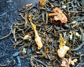 May include: A close-up of loose leaf green tea with dried jasmine flowers. The tea is a mix of dark green and brown leaves with light brown and white flower petals.