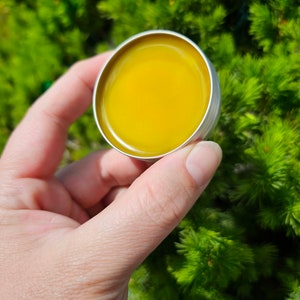 May include: A small round tin container with a yellow balm inside. The tin is held in a hand against a green background.